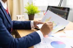 Color photo of torso of seated man in suit holding printouts of charts and graphs in front of a laptop