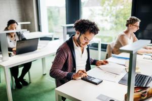 Color photo of bespectacled Black man pressing calculator buttons at his desk in an office