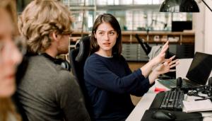 White woman gestures to white man in front of a computer keyboard and laptop as if explaining something
