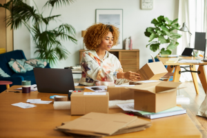 Black woman sits at desk with 3 boxes, a laptop, and papers covering the desk