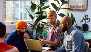 3 men of various races sit in brightly colored clothes and look at a laptop screen