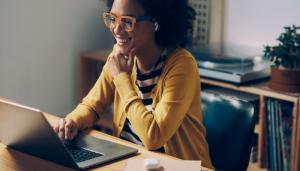 Bespectacled black woman in yellow cardigan smiles at her open laptop screen