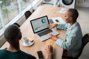 Color overhead shot of Black woman sitting in front of a laptop and showing a financial projection report to a Black man seated beside her