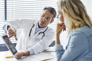 Color photo of older white man in lab coat with a stethoscope shows woman image on iPad