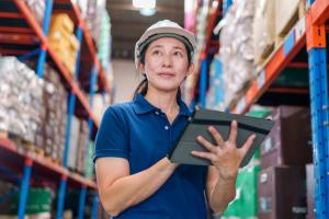 Color photo of female worker in a white hard hat and blue polo and holding a tablet while in a factory