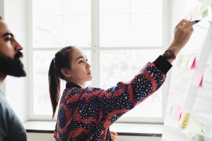 Color photo of young brunette lady in front of a whiteboard covered in Post-It notes with a bearded man beside her