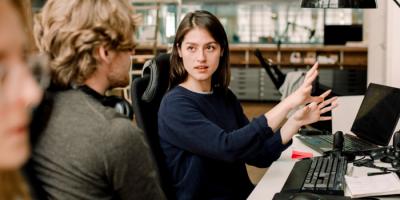 White woman gestures to white man in front of a computer keyboard and laptop as if explaining something