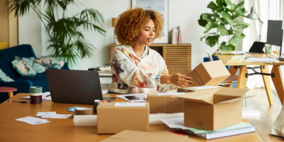 Black woman sits at desk with 3 boxes, a laptop, and papers covering the desk