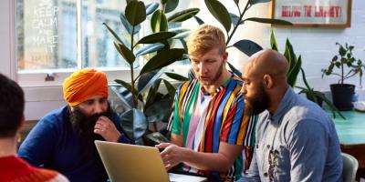 3 men of various races sit in brightly colored clothes and look at a laptop screen