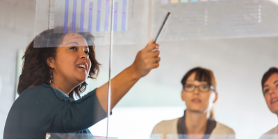 Color photo of woman of color pointing to graphs in front of two white colleagues