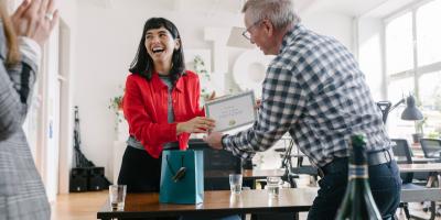 Color photo of smiling white woman in red jacket receiving an award from a coworker at an office