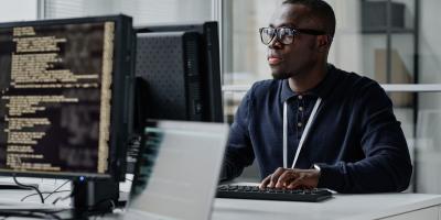 Color photo of young Black man sitting in front of a computer monitor at an office 