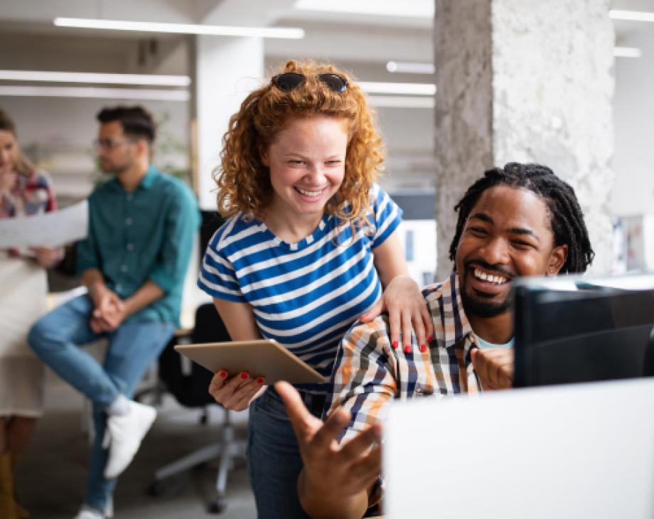 Stock color photo of a red-haired woman laughing with her male colleague in front of a monitor at an office