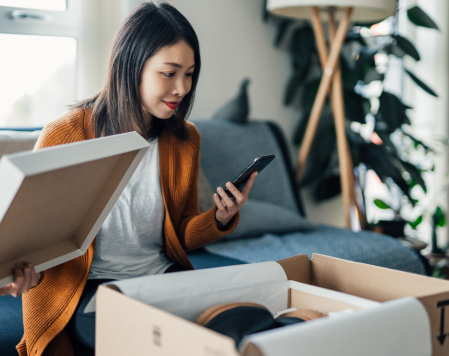 Young Asian woman on couch looks at her mobile phone as she opens a package