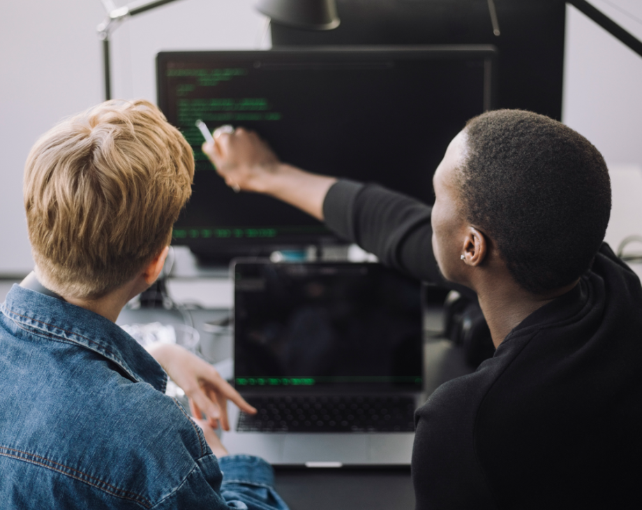 Color photo of Black man seated next to a white man and pointing to a desktop monitor displaying lines of code 
