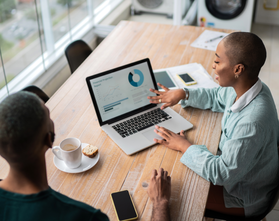 Color overhead shot of Black woman sitting in front of a laptop and showing a financial projection report to a Black man seated beside her