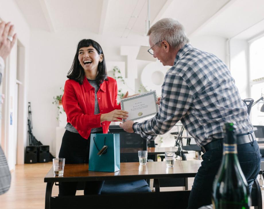 Color photo of smiling white woman in red jacket receiving an award from a coworker at an office