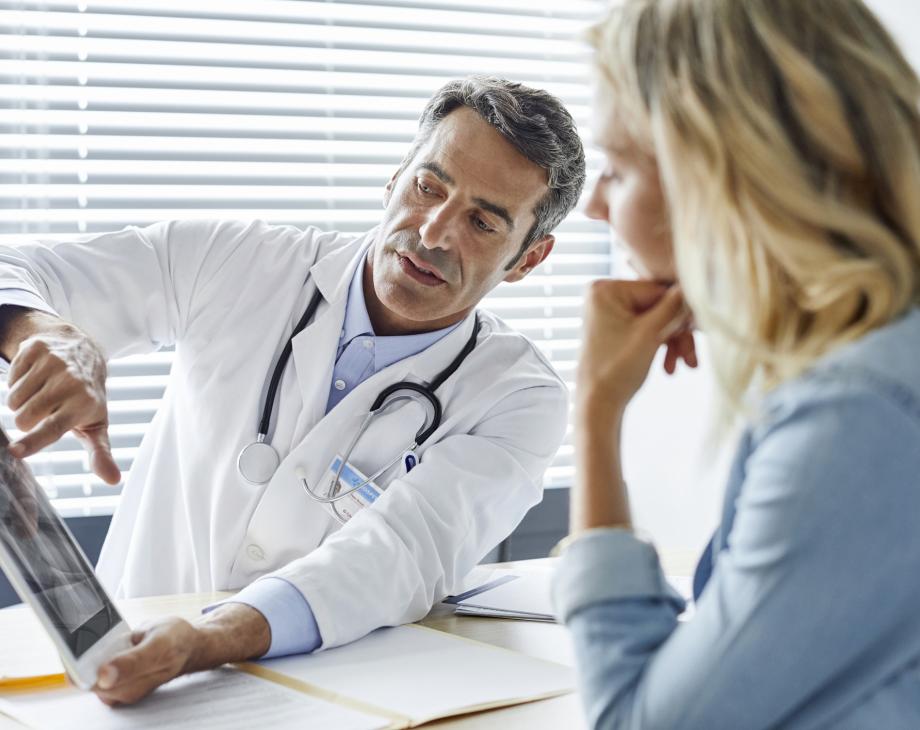 Color photo of older white man in lab coat with a stethoscope shows woman image on iPad