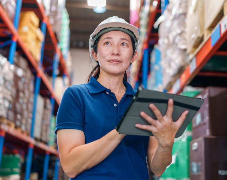 Color photo of female worker in a white hard hat and blue polo and holding a tablet while in a factory