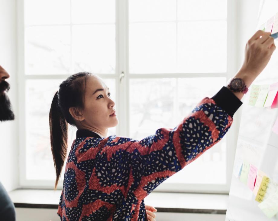 Color photo of young brunette lady in front of a whiteboard covered in Post-It notes with a bearded man beside her