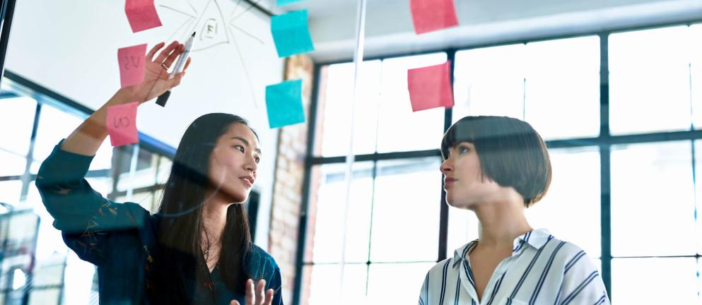 Two women discussing sticky notes on glass wall
